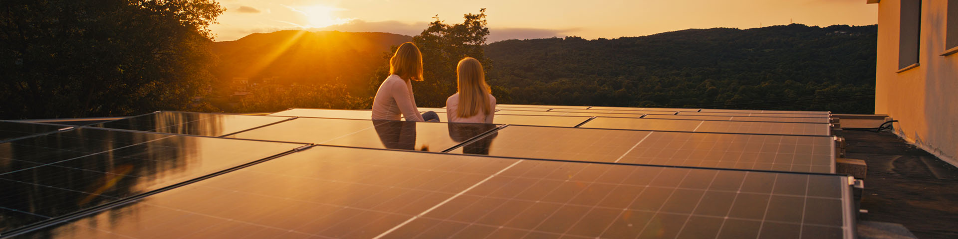 2 personnes assises sur un toit avec des panneaux photovoltaïques regardant un coucher de soleil