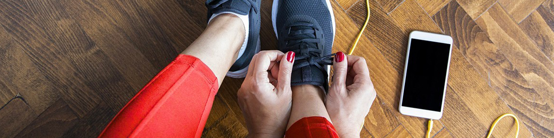 Personne en tenue de sport rouge lace des baskets noires, avec un smartphone et des écouteurs jaunes posés au sol.