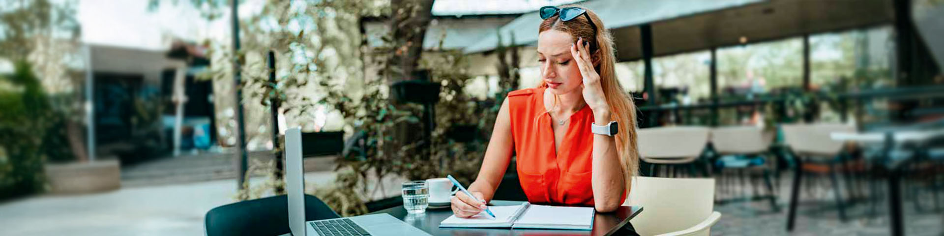 Une jeune femme, lunette de soleil sur la tête, est attablée à une terrasse de café devant son ordinateur portable et en prenant des notes.