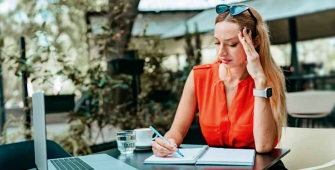 Une jeune femme, lunette de soleil sur la tête, est attablée à une terrasse de café devant son ordinateur portable et en prenant des notes.