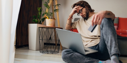 Un homme la main sur le front qui regarde son ordinateur avec sa carte bleue dans la main gauche